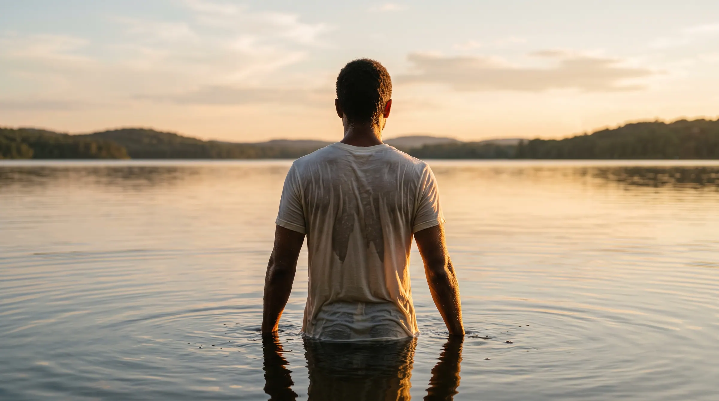 A man standing waist-deep in still water at golden hour, wearing a white tee